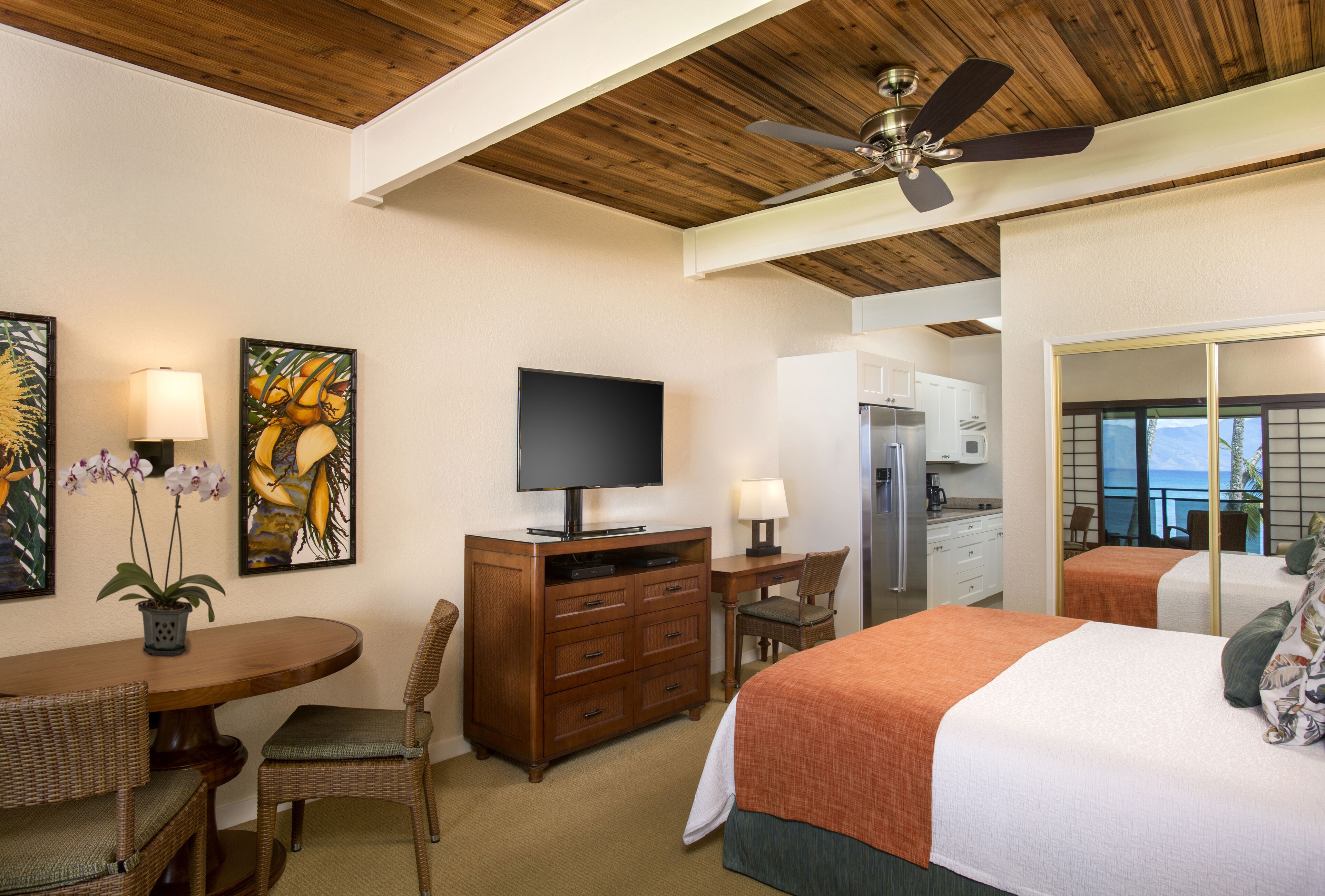 A hotel room with a wooden ceiling, a flat-screen TV, a dining table with chairs, a bed with an orange blanket, and a view of the ocean through a large window.