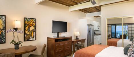 A hotel room with a wooden ceiling, a flat-screen TV, a dining table with chairs, a bed with an orange blanket, and a view of the ocean through a large window.