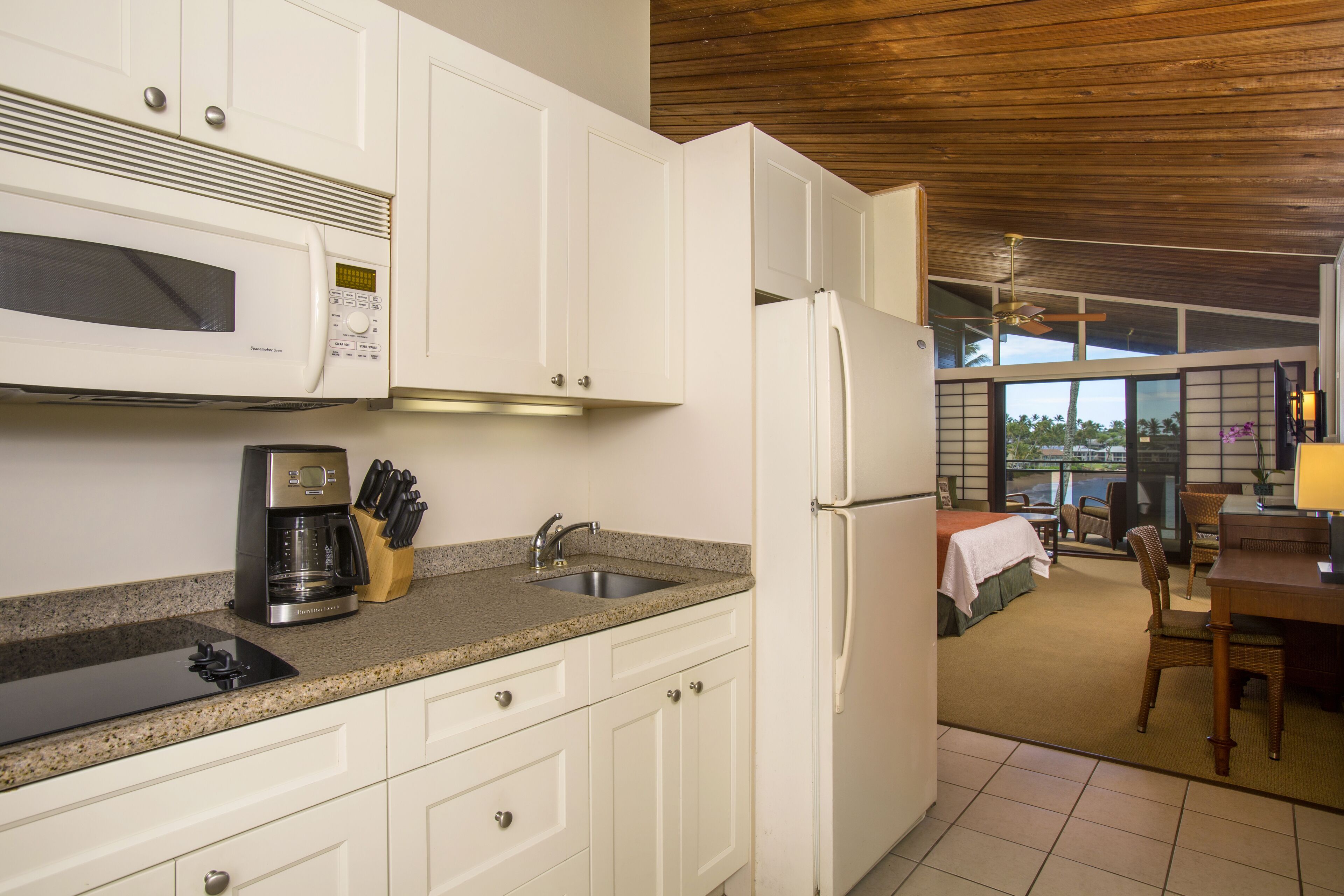 Una cocina con armarios blancos, microondas, cafetera y refrigerador. Hay un comedor con mesa y sillas.
