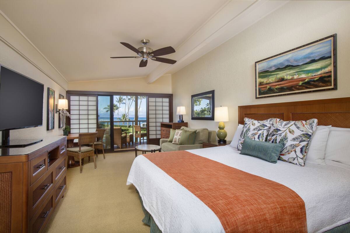 Quarto de hotel com cama, televisão, ventoinha no teto, quadro e vista para o exterior.