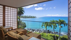 A balcony with wicker furniture, a glass table, and a view of the ocean and a tropical island. - Napili Kai Beach Resort (Lahaina)