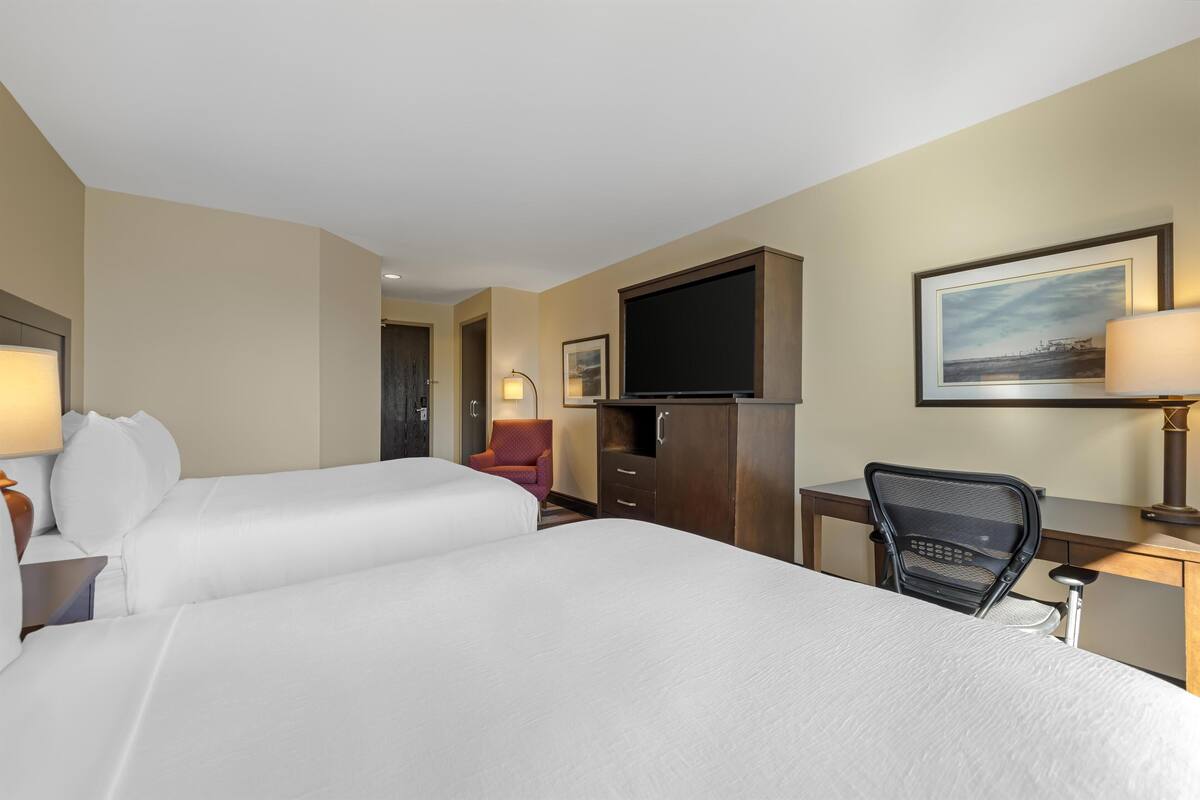 A hotel room with two beds, a desk, a chair, a TV, and a lamp.
