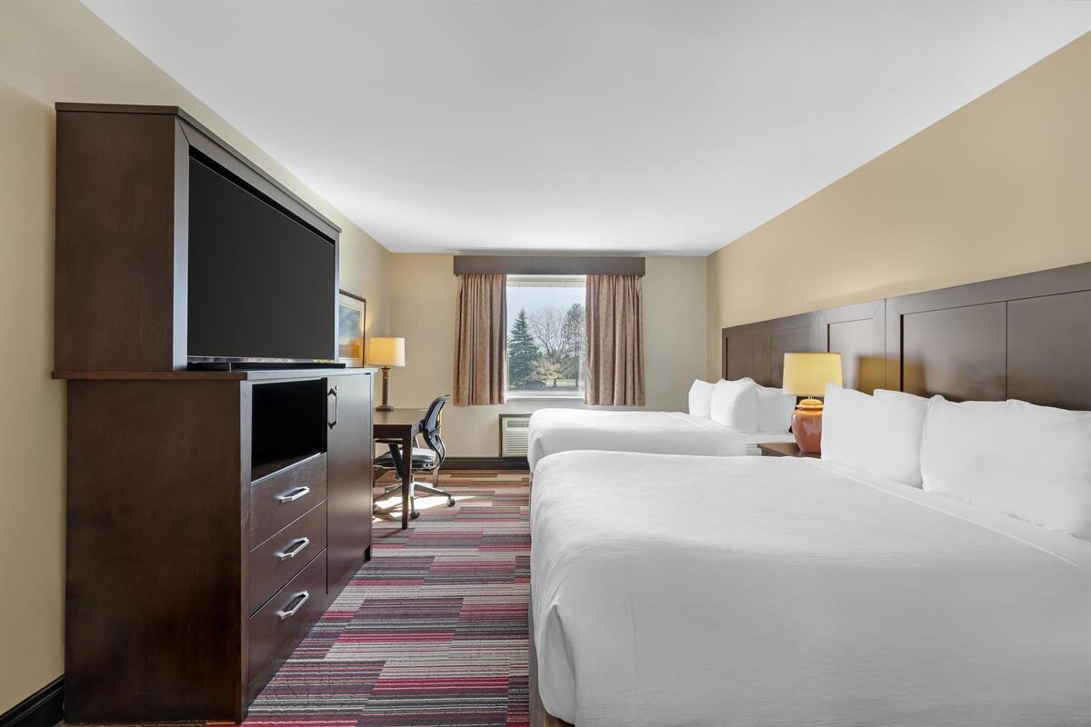A hotel room with two beds, a desk, a TV, and a window with curtains.