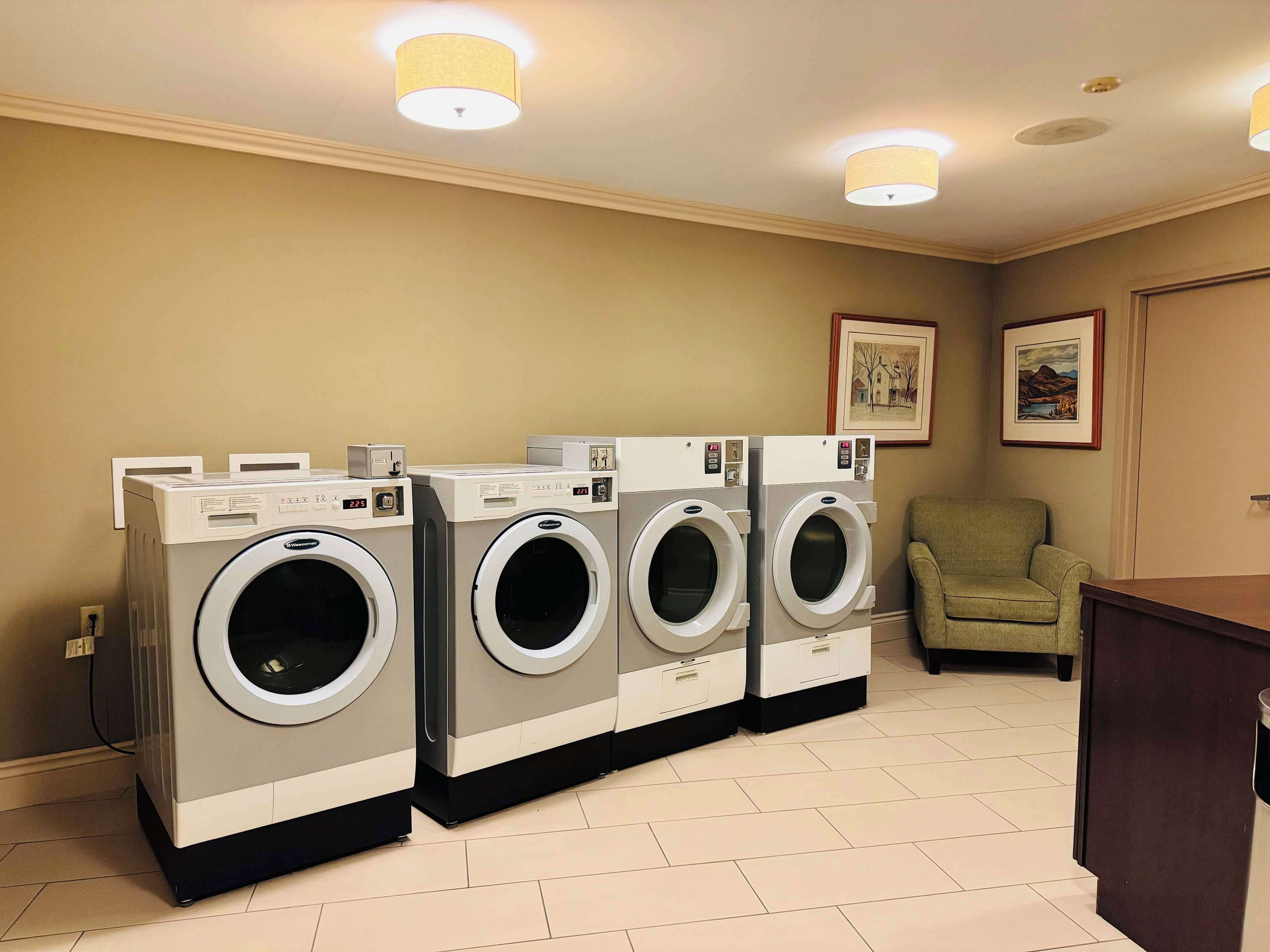 A laundry room with four front-loading washing machines, a chair, and framed artwork on the wall.