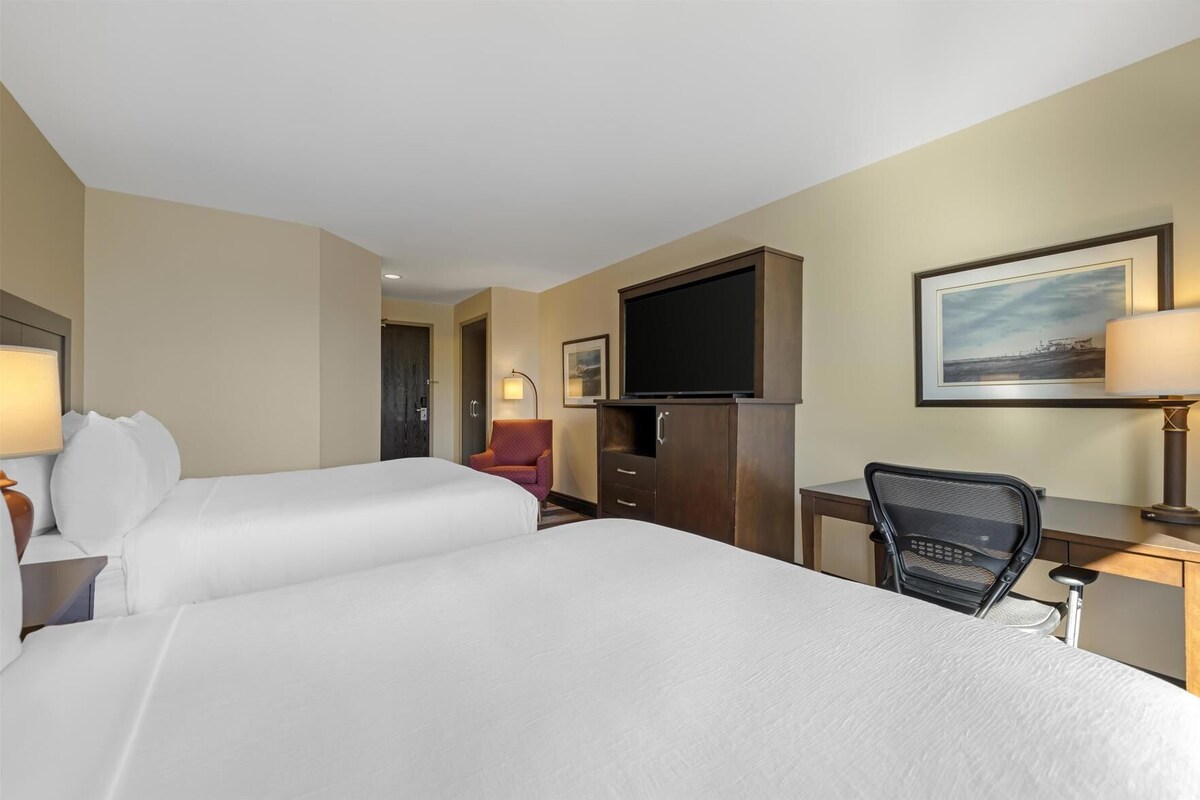 A hotel room with two beds, a desk, a chair, a television, and a painting on the wall.
