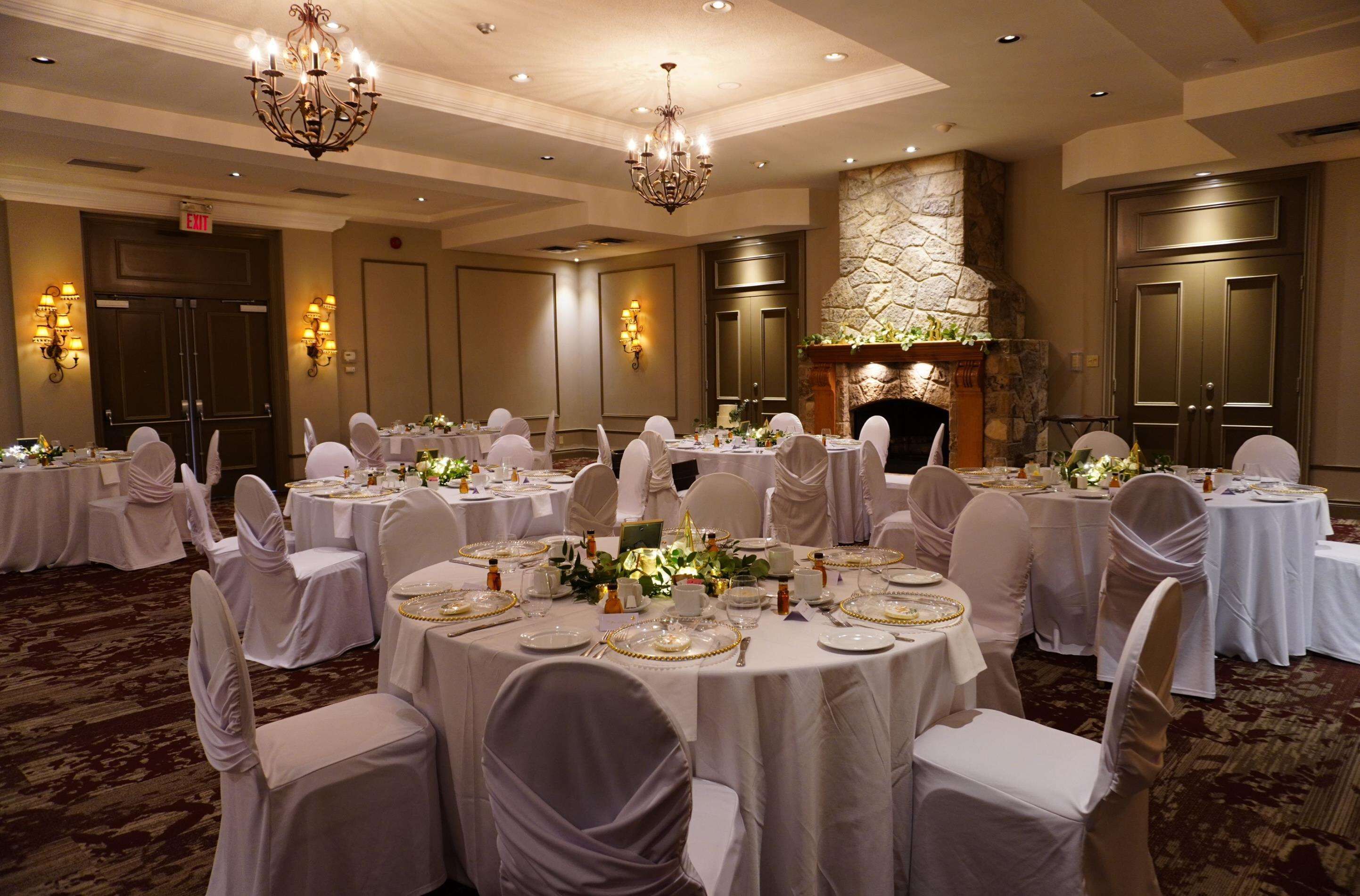 Un salón de banquetes con mesas redondas dispuestas para un evento formal, con candelabros, chimenea de piedra y una pared de piedra.