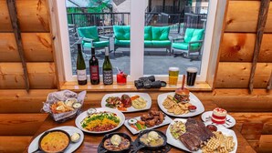 A dining table with various dishes, drinks, and a camera on a wooden bench by a window with a view of green outdoor seating.