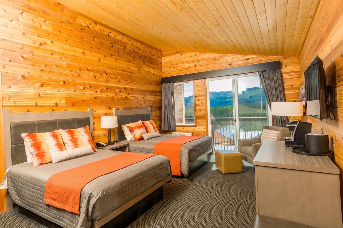 A log cabin-style room with two beds, a desk, and a large window offering a mountain view.