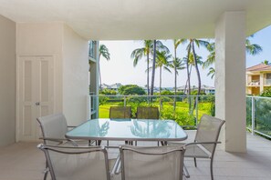 A balcony with a glass table and four chairs, overlooking a lush garden with palm trees and a distant building. - Vista Waikoloa (Waikoloa)