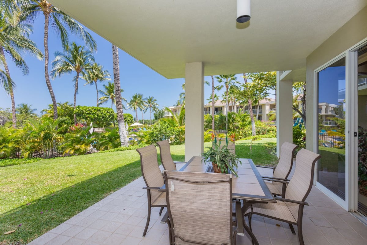 A covered outdoor patio with a table and chairs, surrounded by palm trees and a well-maintained lawn.