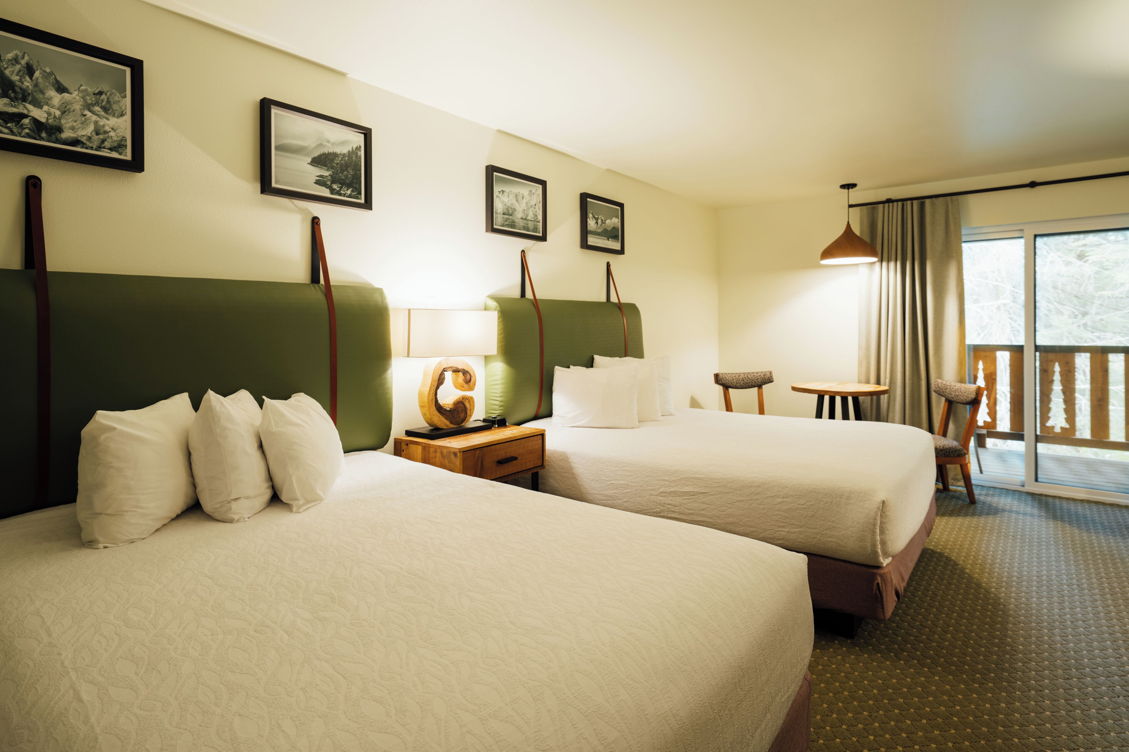 A hotel room with two beds, a small table, a chair, and a balcony with a view.