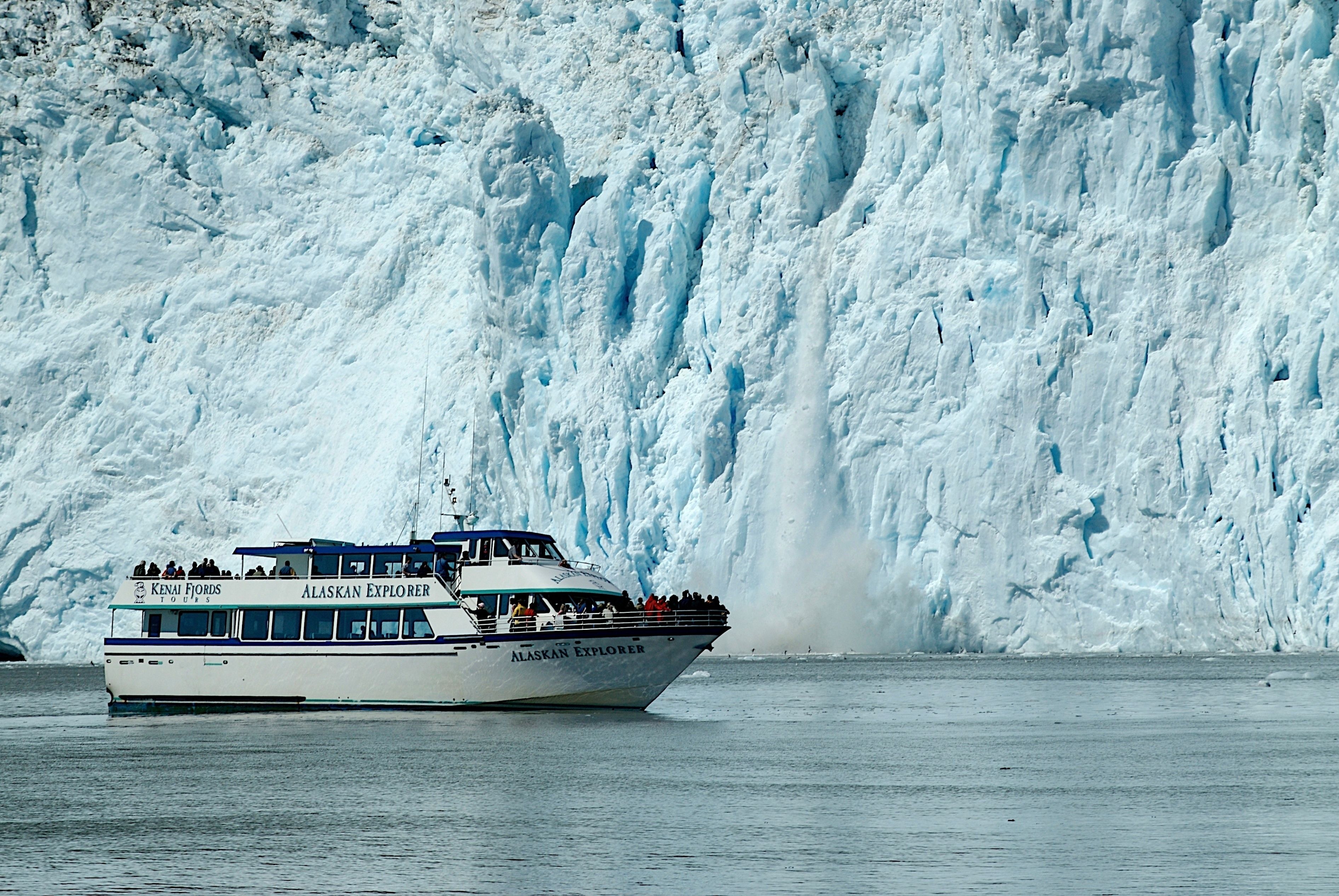 A boat named 'Alaskan Explorer' with passengers, sailing near a large glacier.