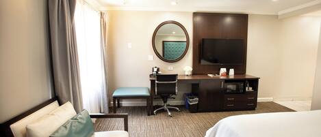 Habitación de hotel con cama, escritorio con silla, televisión, espejo y ventana con cortinas.