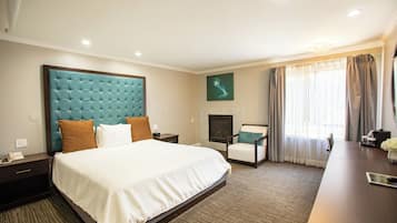 A hotel room with a large bed, a desk, a chair, a fireplace, and a large window with curtains.