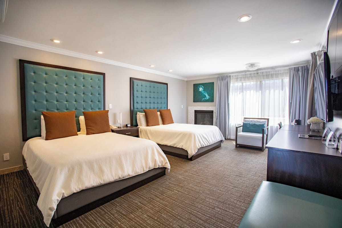 A hotel room with two beds, a desk, a chair, a fireplace, and a large window with curtains.
