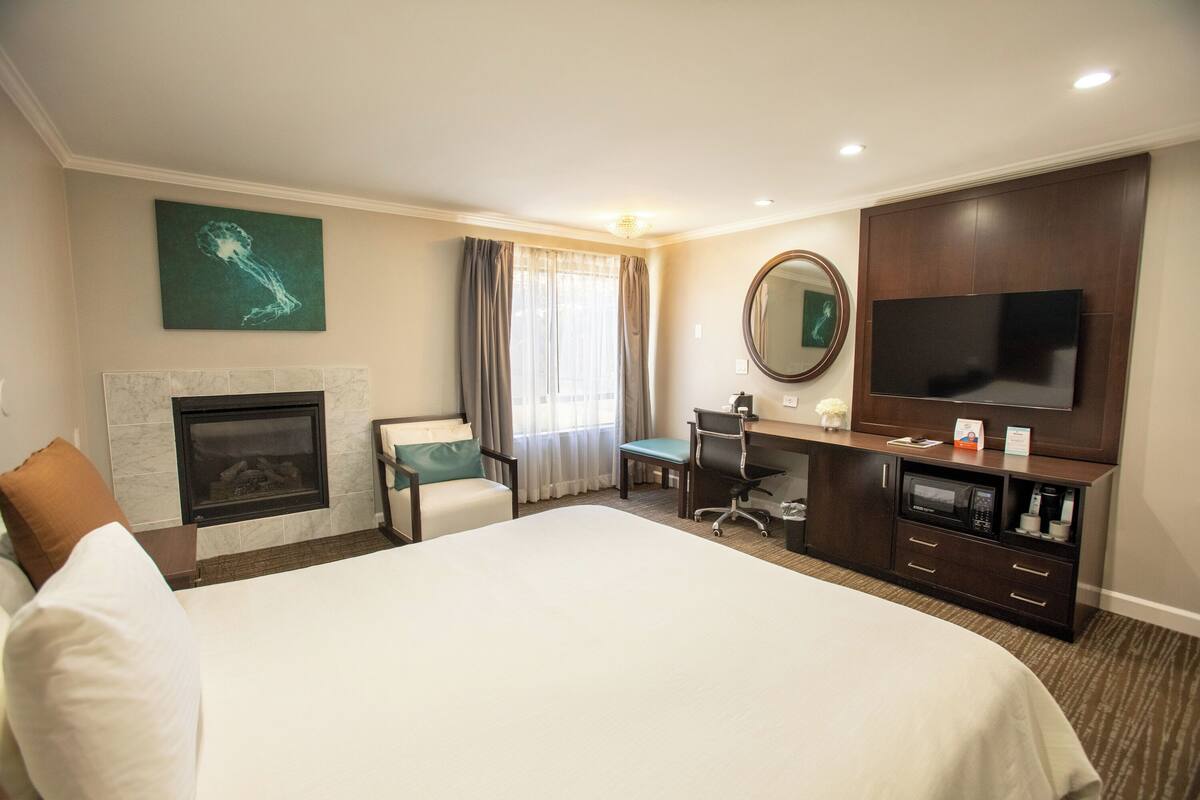 A hotel room with a large bed, a desk with a chair, a television, a fireplace, and a large mirror.
