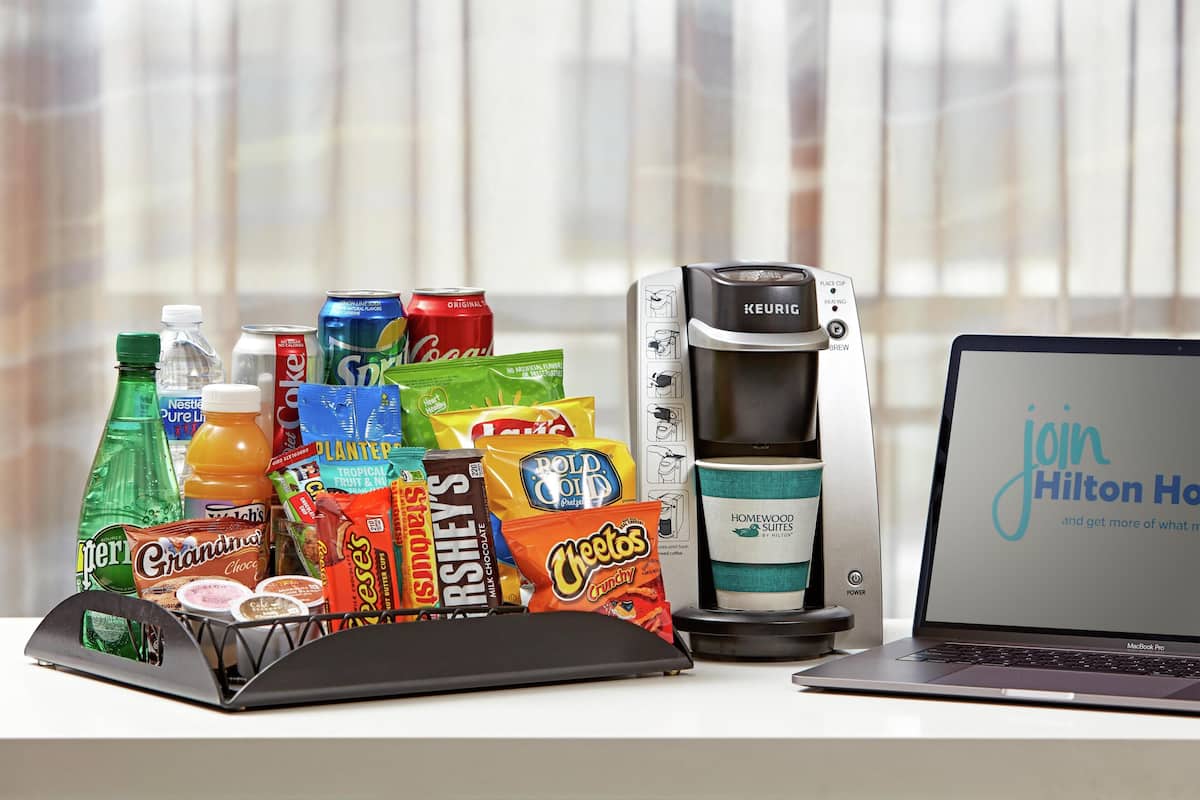 Una cafetera, una computadora portátil y diversos bocadillos sobre una mesa.