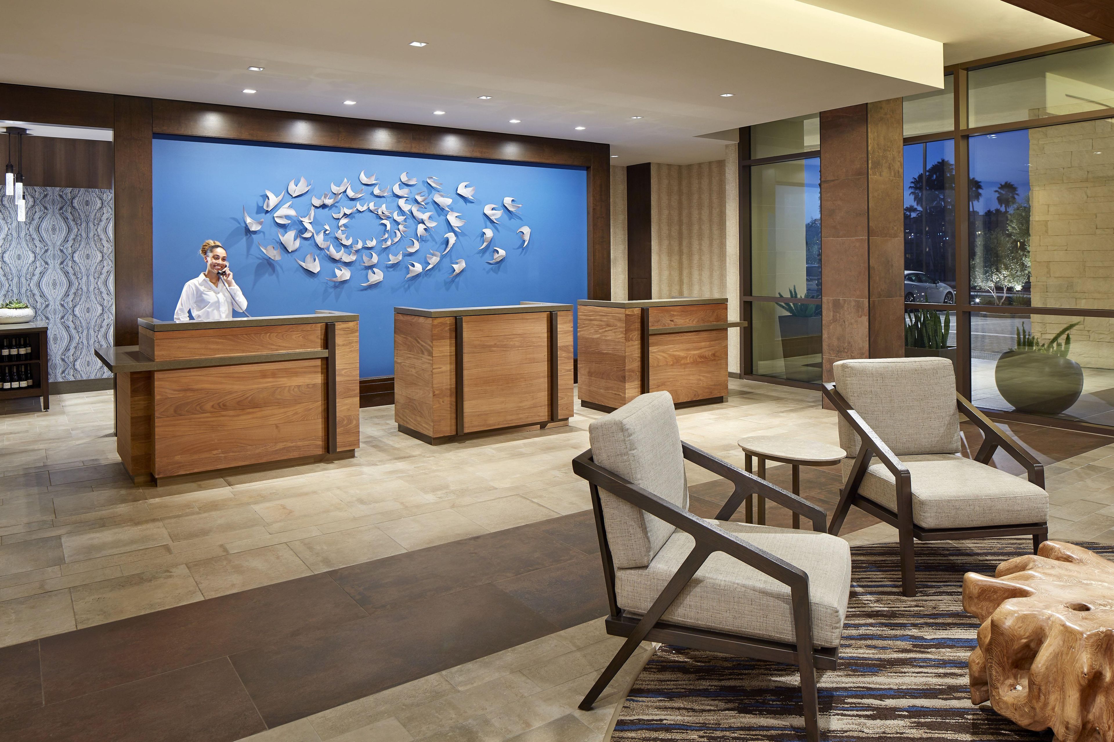 A hotel reception area with a receptionist, wooden reception desks, and a large wall art installation.