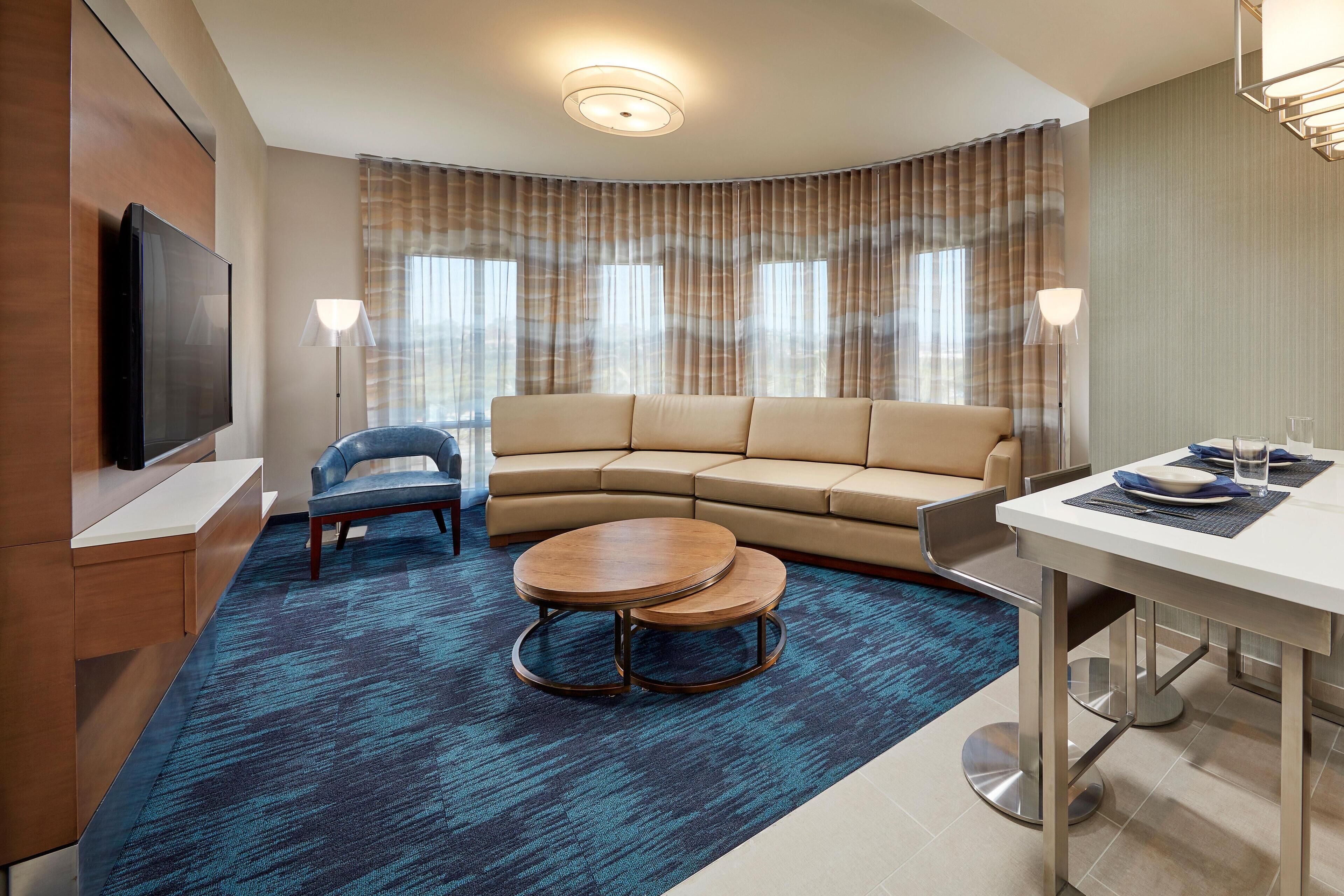 A modern hotel room with a curved sofa, a round coffee table, a flat-screen TV, and a dining area with a table and chairs.
