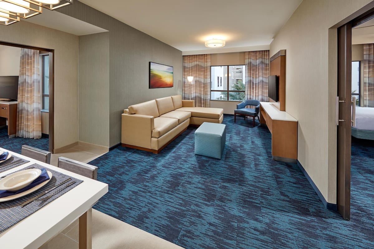 Habitación de hotel con sofá, escritorio, televisión y comedor con una mesa lista para dos personas.