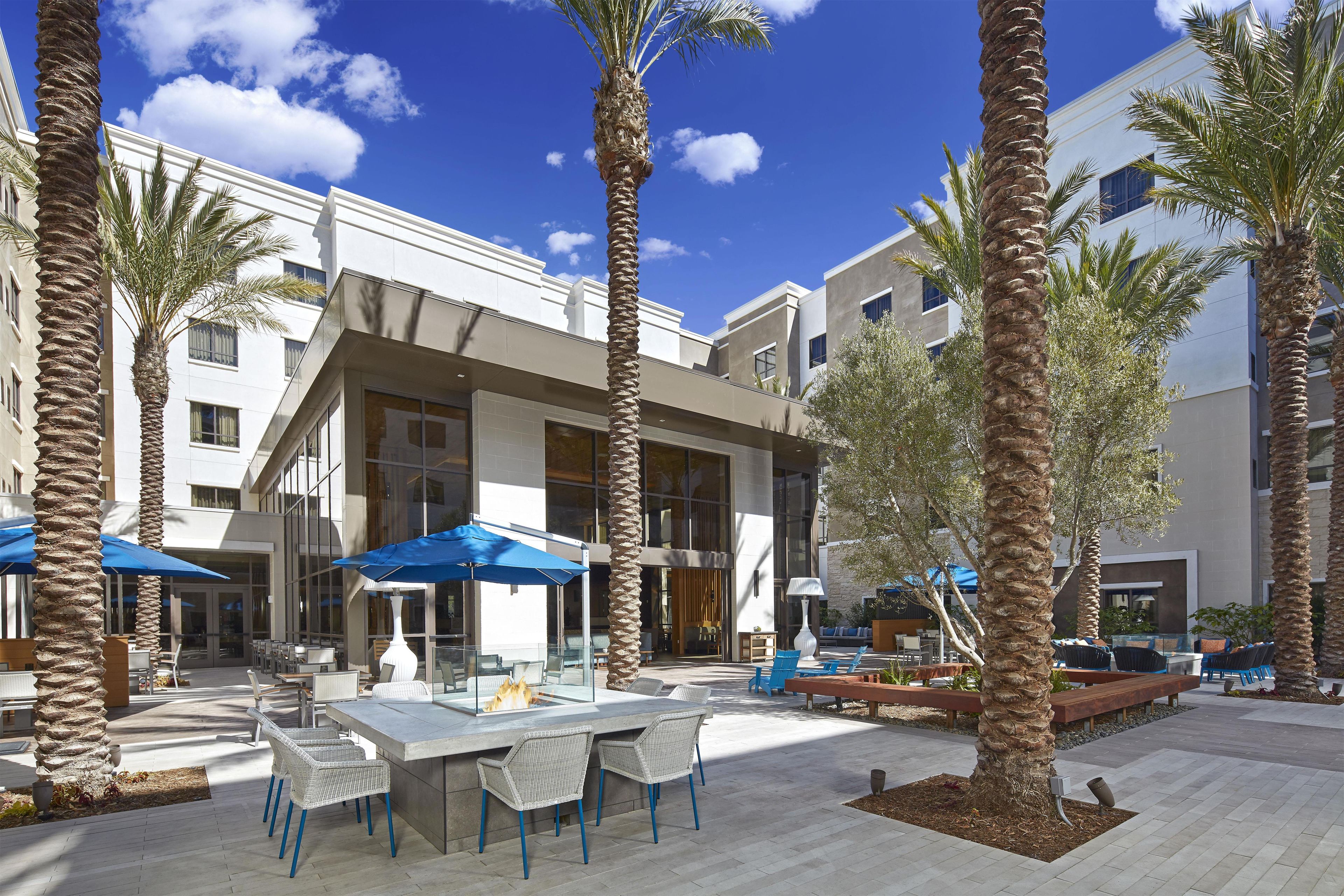 An outdoor patio area with tables and chairs, palm trees, and a modern building with large windows.
