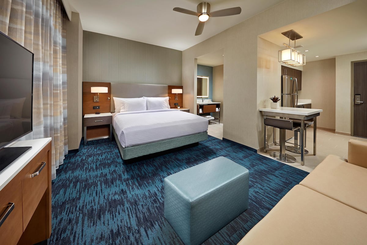 Habitación de hotel con cama, escritorio, silla, televisión, ventilador de techo y baño con espejo y lavamanos.