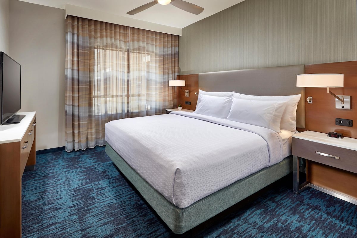 Habitación de hotel con cama, escritorio, televisor, mesita de noche, lámpara y ventilador de techo.