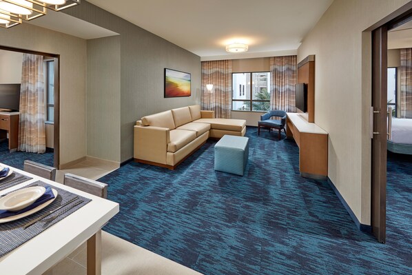 Habitación de hotel con sofá, escritorio, televisión y comedor con una mesa lista para dos personas.