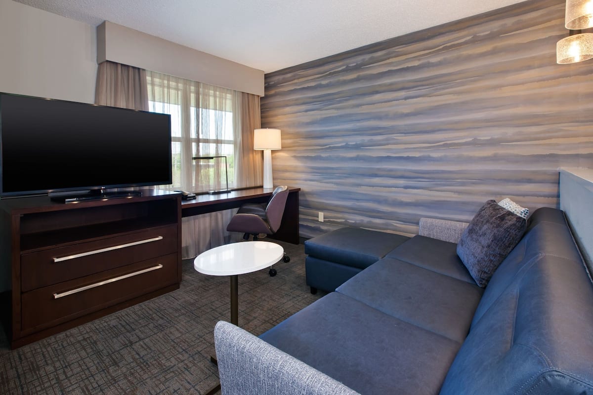 A modern hotel room with a flat-screen TV, a desk with a lamp, a sofa, and a patterned wall.