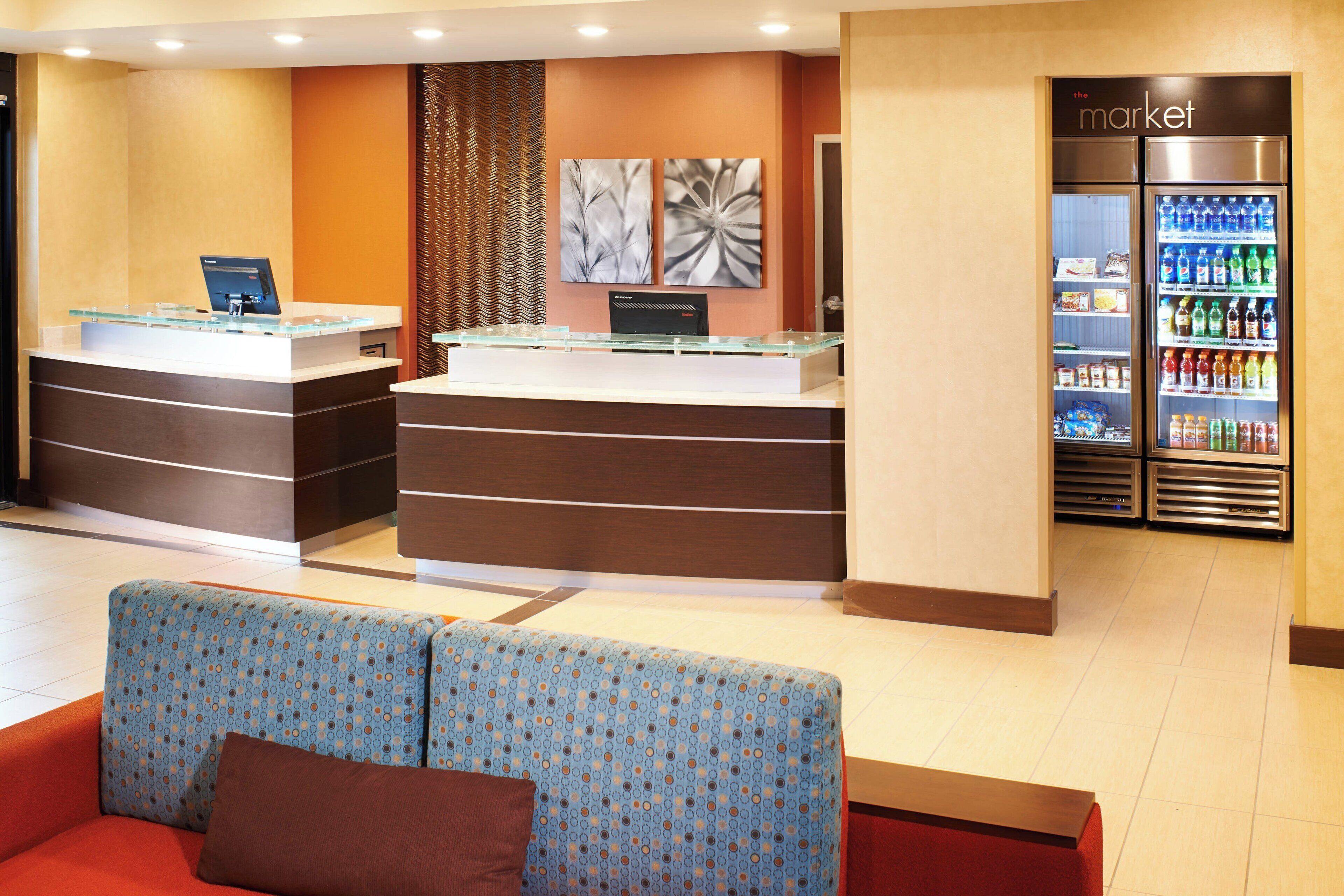 A hotel reception area with a glass counter, a computer, and a refrigerator stocked with beverages. There are two seating areas with patterned upholstery.