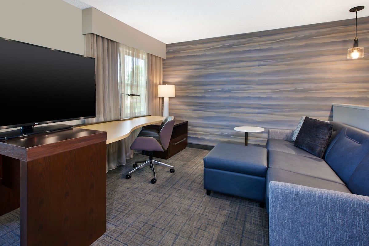 A modern hotel room with a flat-screen TV, a desk with a lamp, a sofa, and a patterned wall.