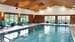An indoor swimming pool with clear blue water, surrounded by blue chairs and tables, wooden ceiling beams, and large windows offering a view of greenery outside. - Residence Inn by Marriott Ann Arbor North (Ann Arbor)