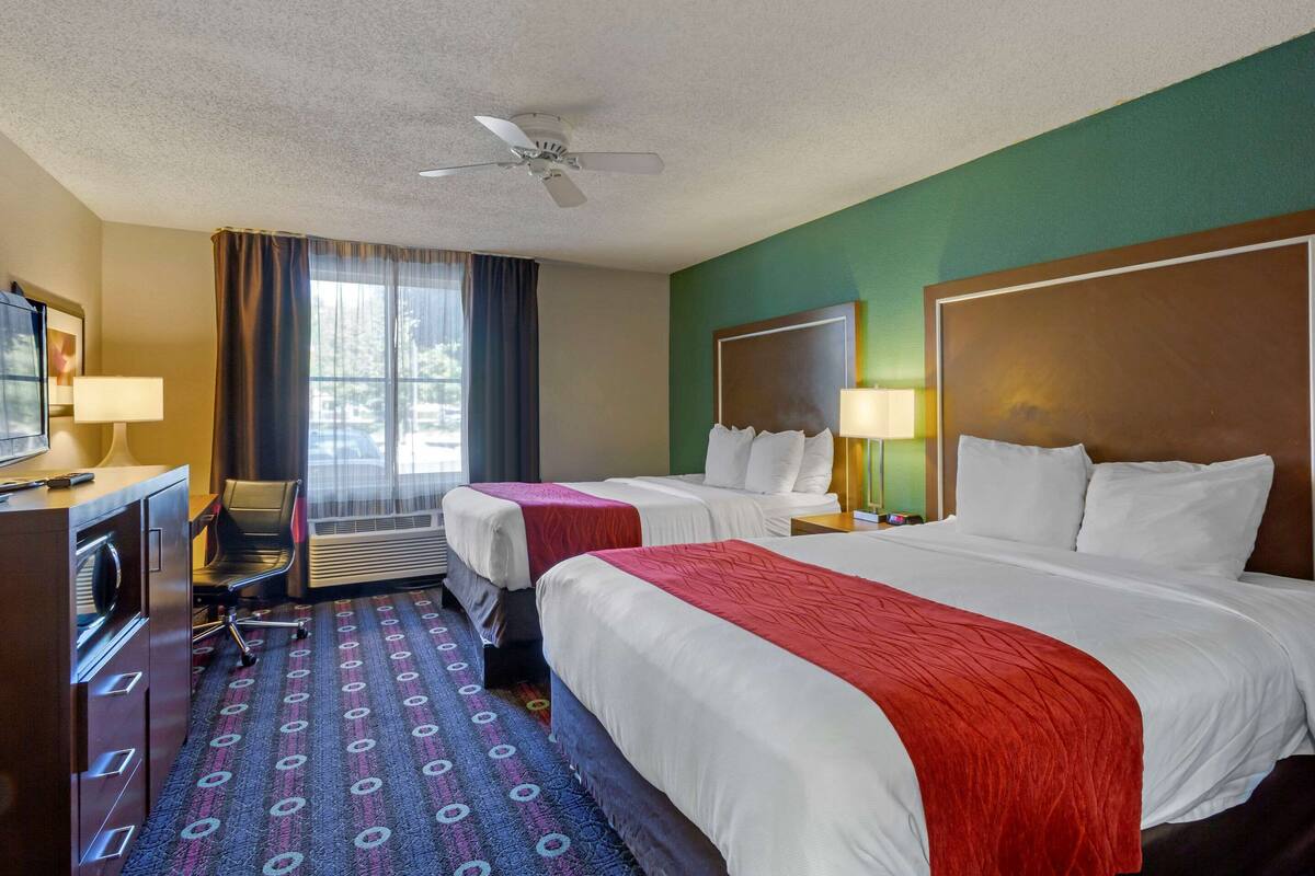 Ein Hotelzimmer mit zwei Betten, einem Schreibtisch mit Stuhl, einem Deckenventilator und einem großen Fenster mit Vorhängen.