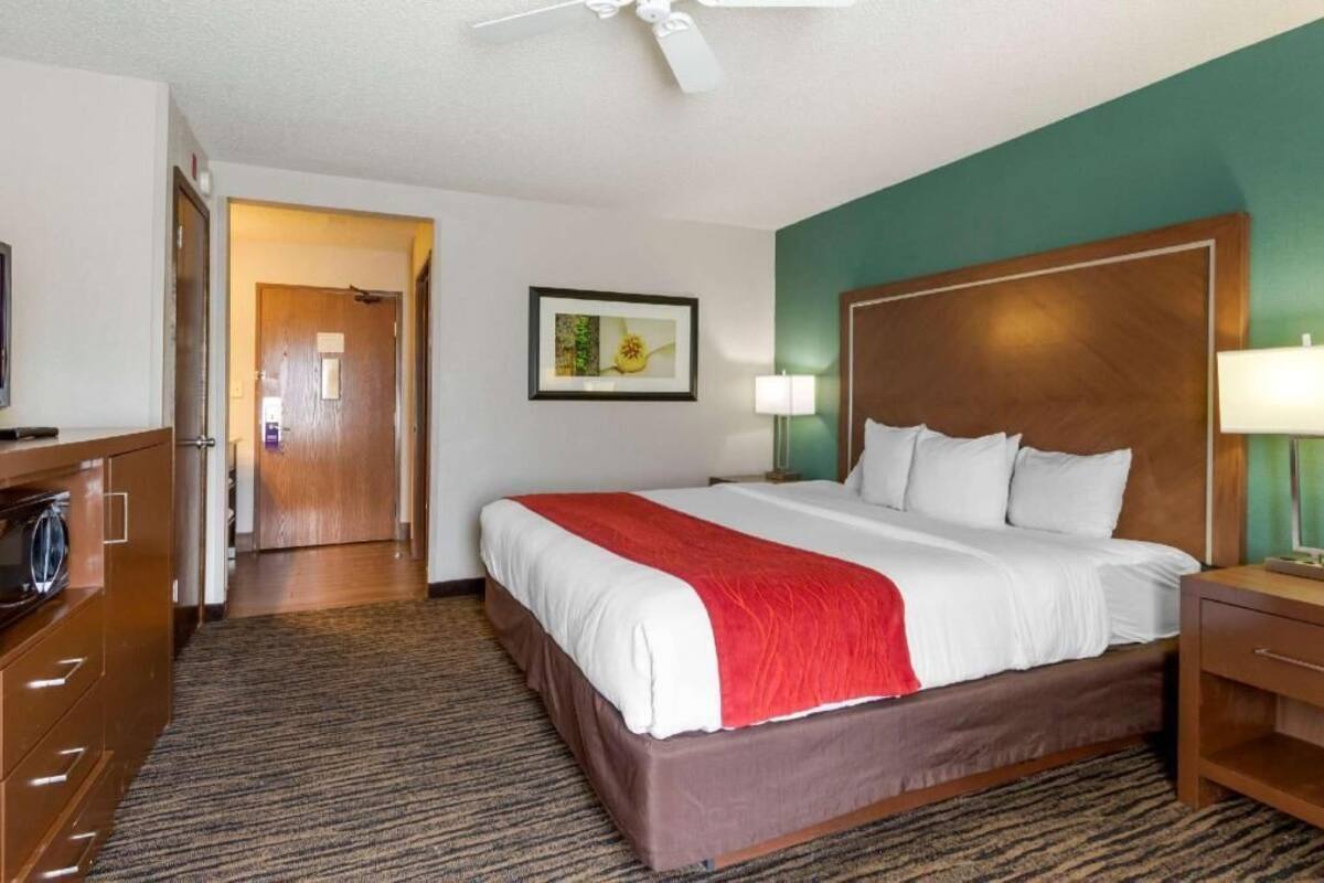 Ein Hotelzimmer mit einem Bett, einem Schreibtisch, einem Sessel, einer Lampe, einem Deckenventilator und einer Tür zu einem weiteren Raum.