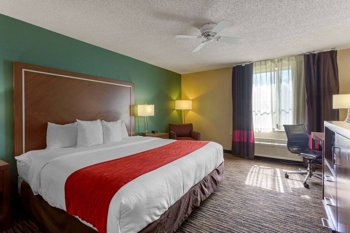 Ein Hotelzimmer mit einem großen Bett, zwei Nachttischlampen, einem Schreibtisch mit Stuhl, einem Deckenventilator und Blick ins Freie durch ein Fenster mit Vorhängen.