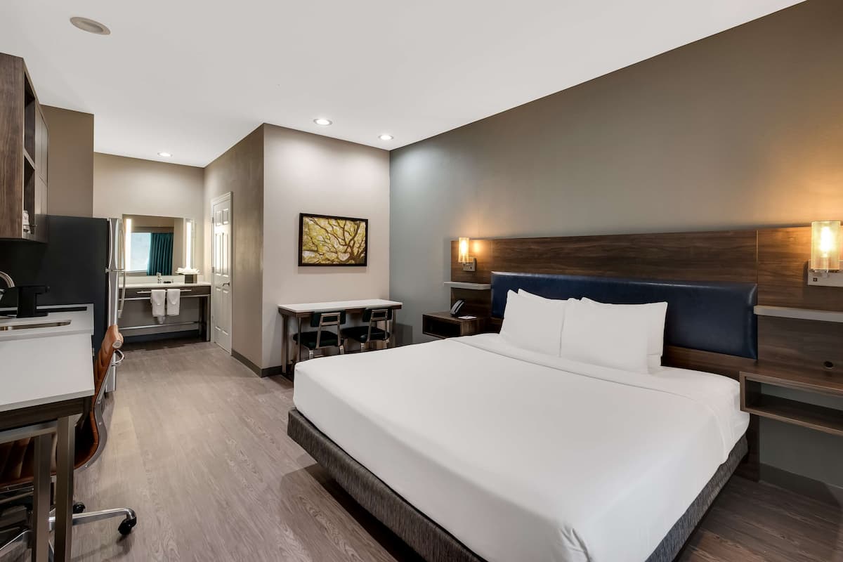A modern hotel room with a large bed, a desk, a chair, a vanity with a mirror, and a wall-mounted light fixture.
