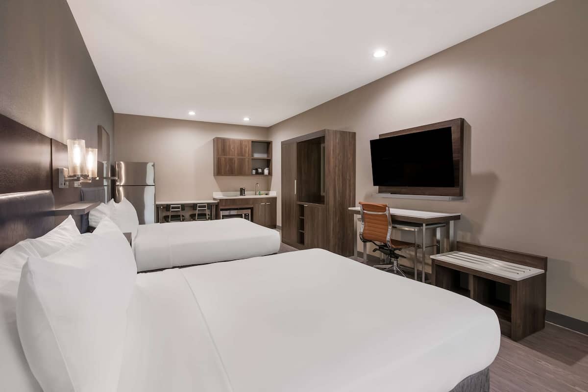 A modern hotel room with two beds, a desk, a chair, a TV, and a kitchenette.