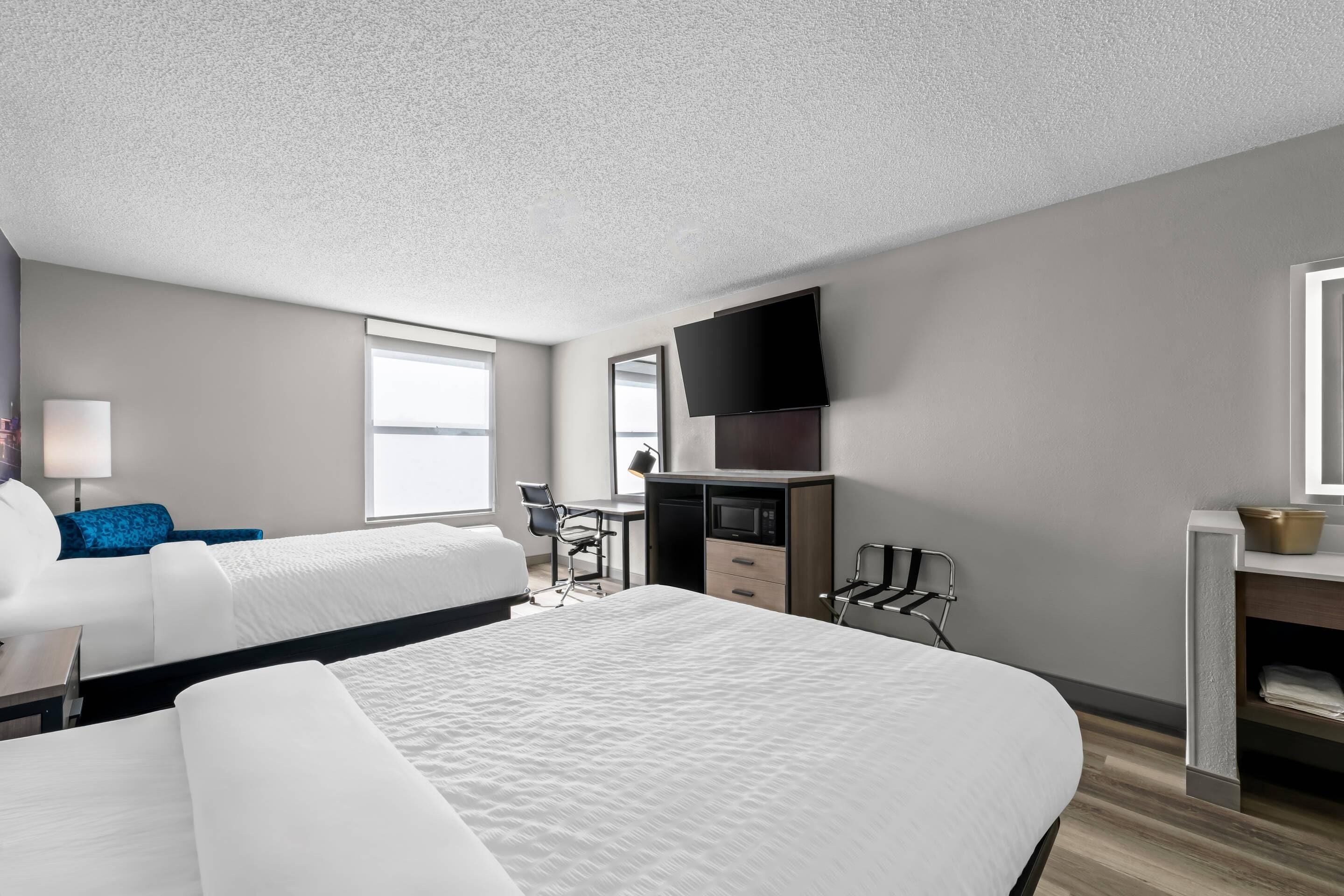 A modern hotel room with a large bed, a desk with a chair, a flat-screen TV mounted on the wall, and a window providing natural light.