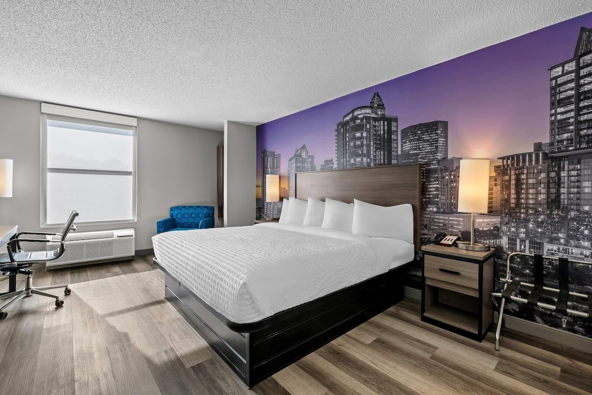 A modern hotel room with a large bed, a cityscape mural, a desk with a lamp, and a chair.