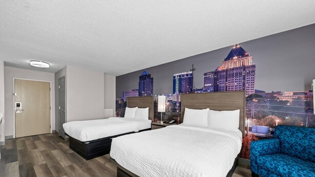 A hotel room with two beds, a cityscape mural, a blue sofa, and a wooden floor. - Clarion Pointe (High Point)