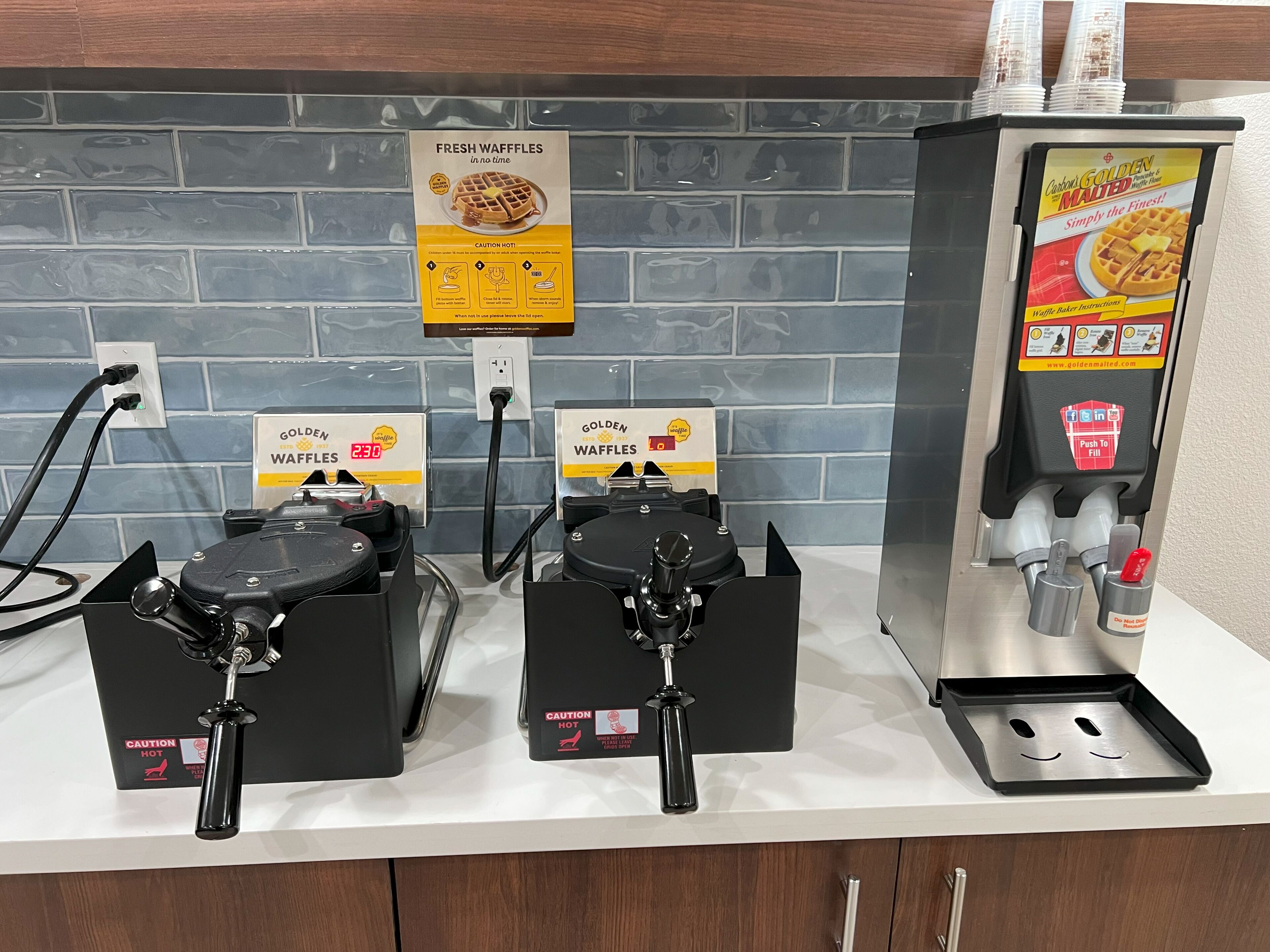 A waffle-making station with two waffle makers, a sign for Golden Waffles, and a push-to-fill dispenser.