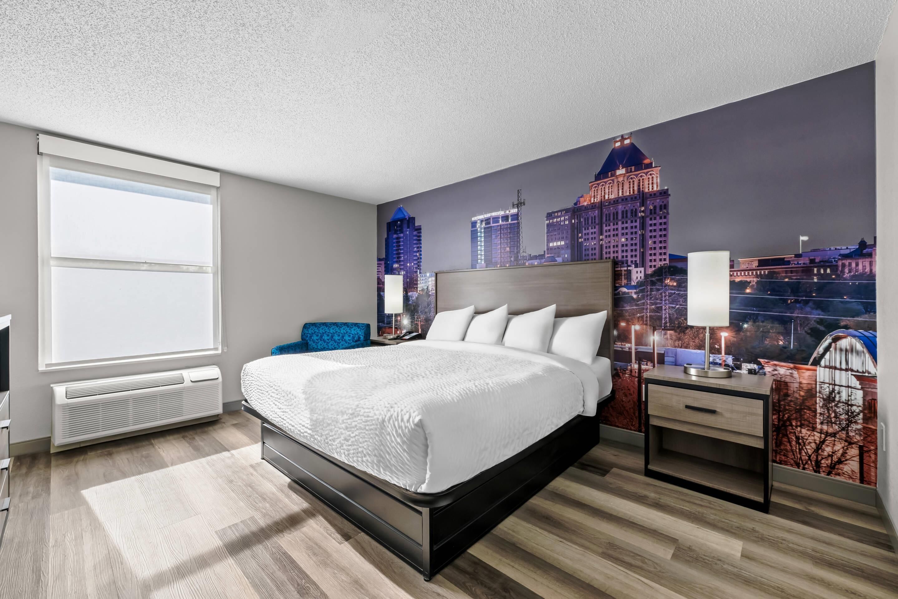 A hotel room with a large bed, a nightstand, a desk, and a cityscape mural on the wall.