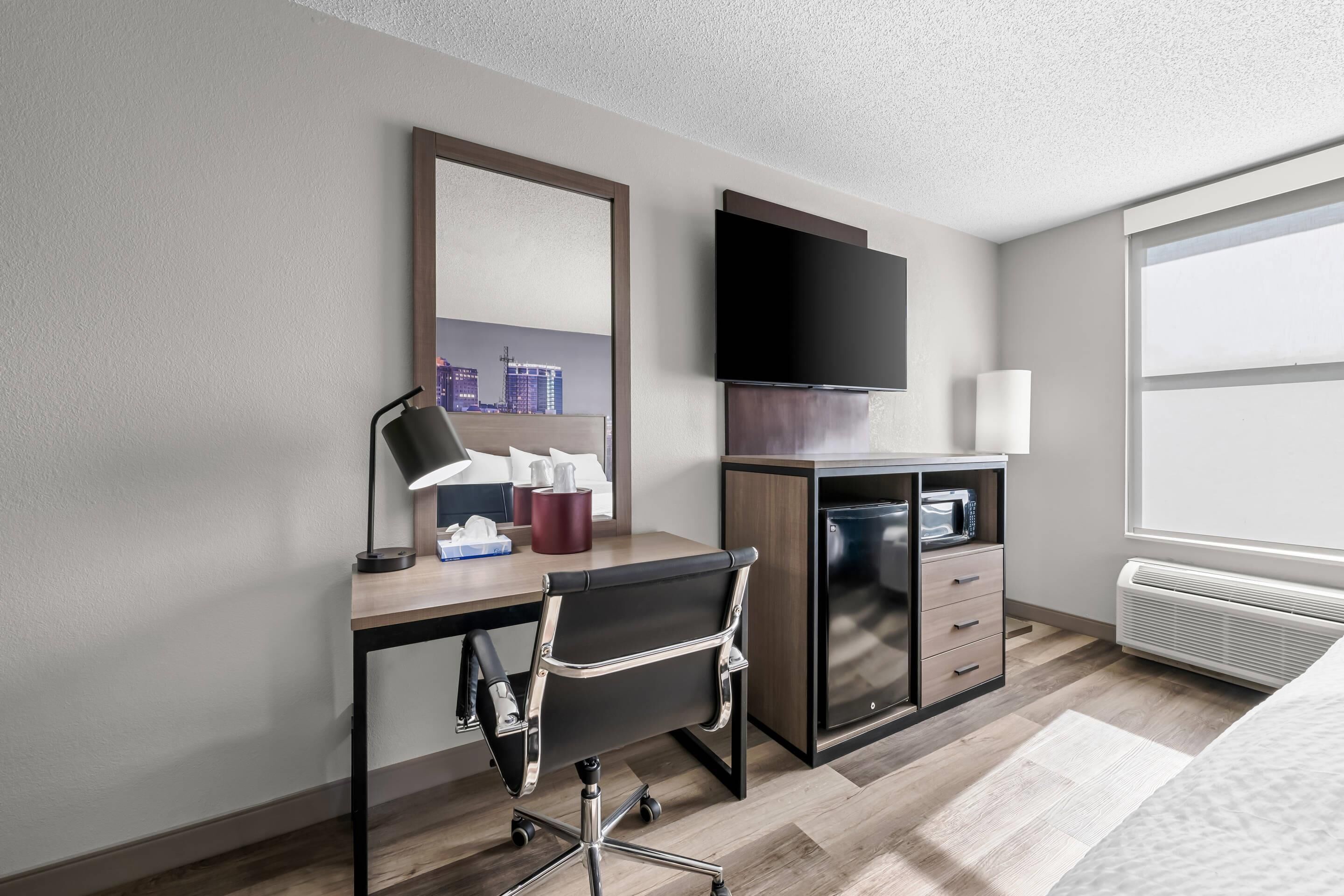 A modern hotel room with a wooden desk, a black office chair, a flat-screen TV, a microwave, and a large window with a city view.