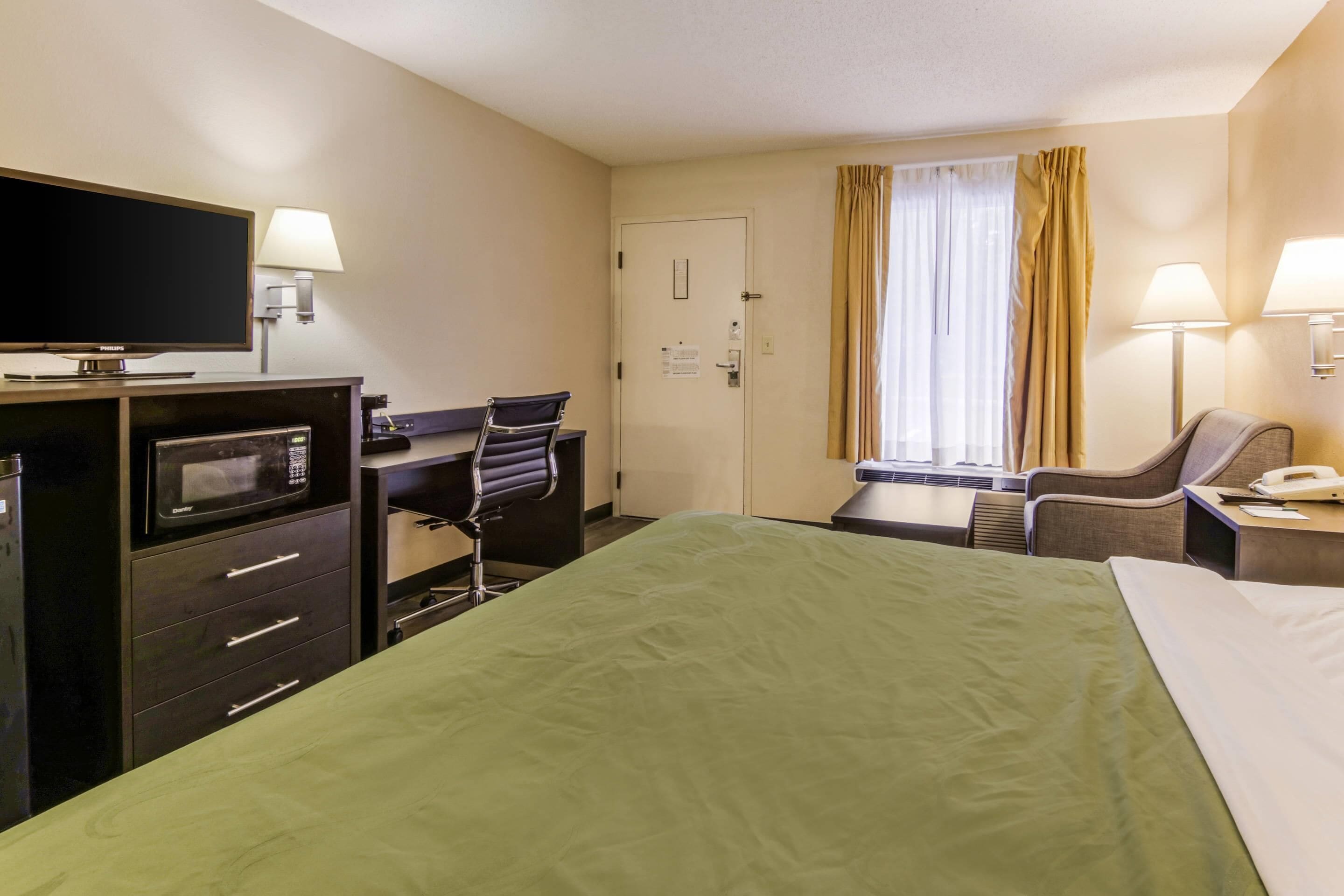 A hotel room with a bed, a desk with a computer, a chair, a TV, a microwave, and a window with curtains.