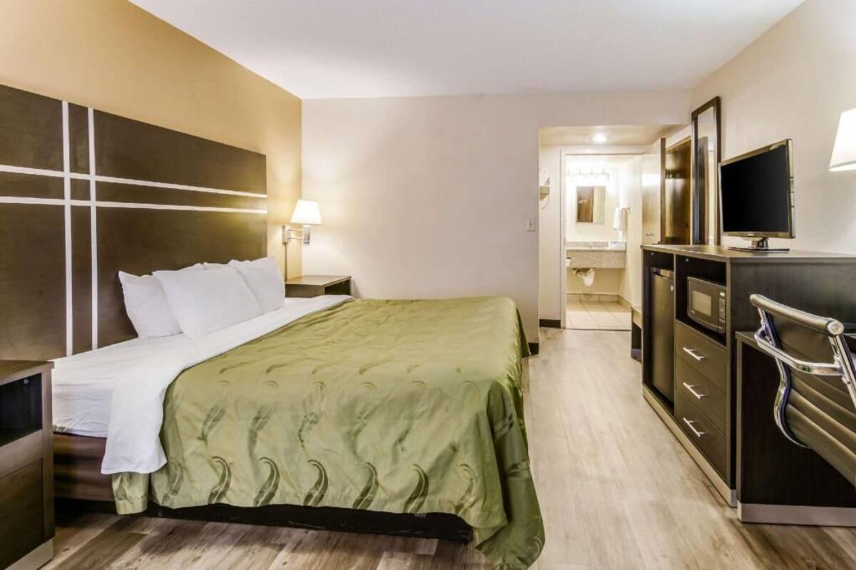 A hotel room with a bed, bedside table, lamp, TV, and a dresser.