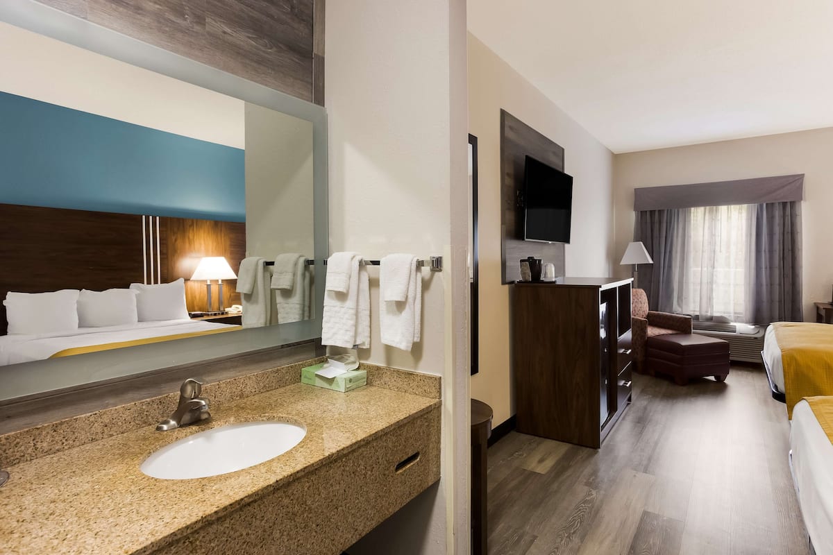 Une chambre d’hôtel avec un lavabo doté d’un plan de travail en granit, un grand miroir, un lit avec une tête de lit jaune, un téléviseur fixé au mur et une fenêtre avec des rideaux.