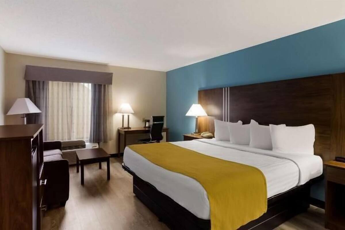 Une chambre d’hôtel avec un lit, des tables de chevet, un bureau, une chaise et une fenêtre avec des rideaux.