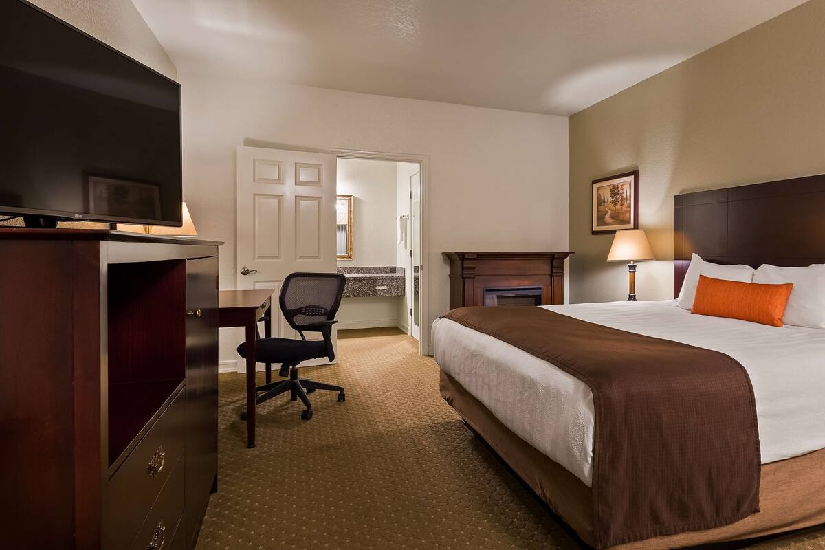 Quarto de hotel com cama, escrivaninha com cadeira, televisão, lareira e um quadro na parede.