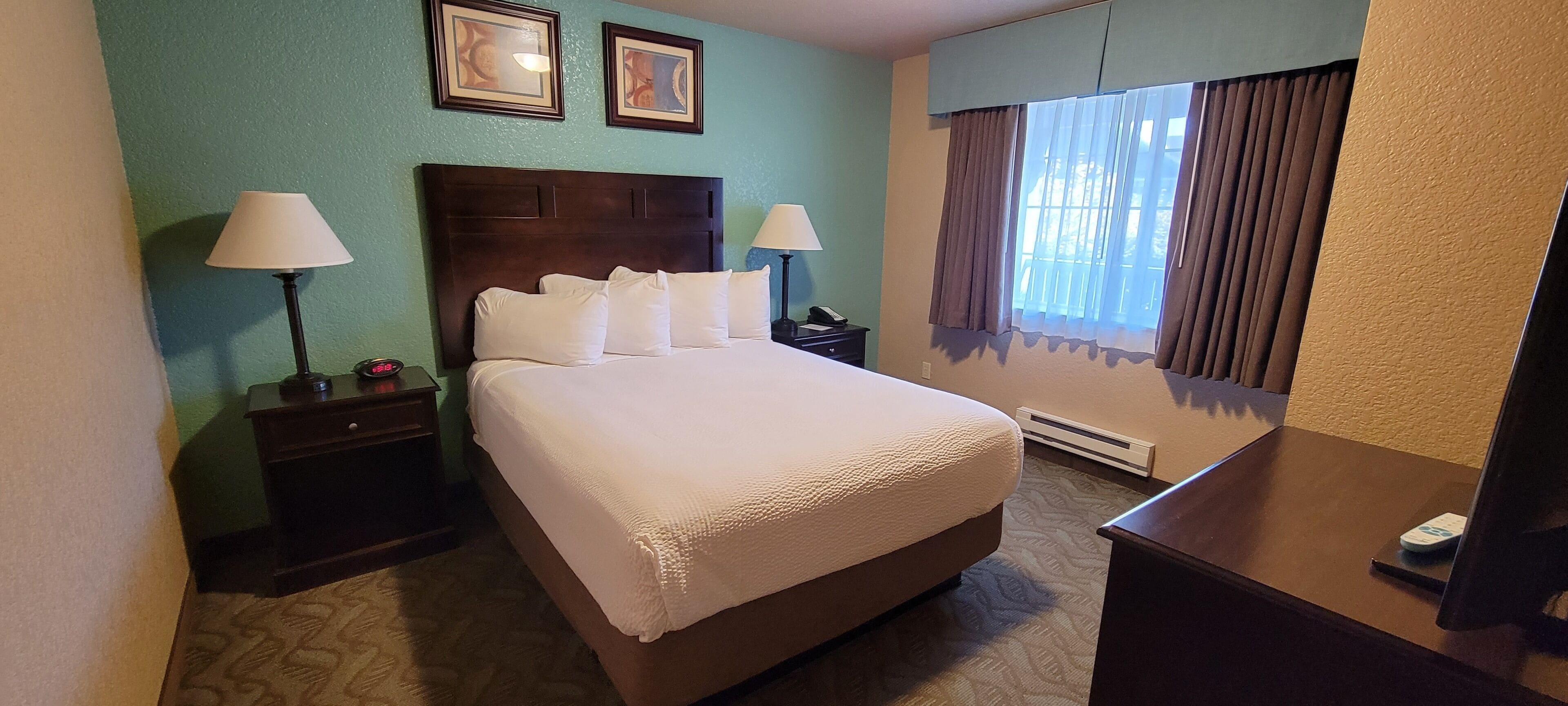 Habitación de hotel con una cama grande, dos mesitas de noche con lámparas, un mueble auxiliar, una ventana con cortinas y dos cuadros en la pared.