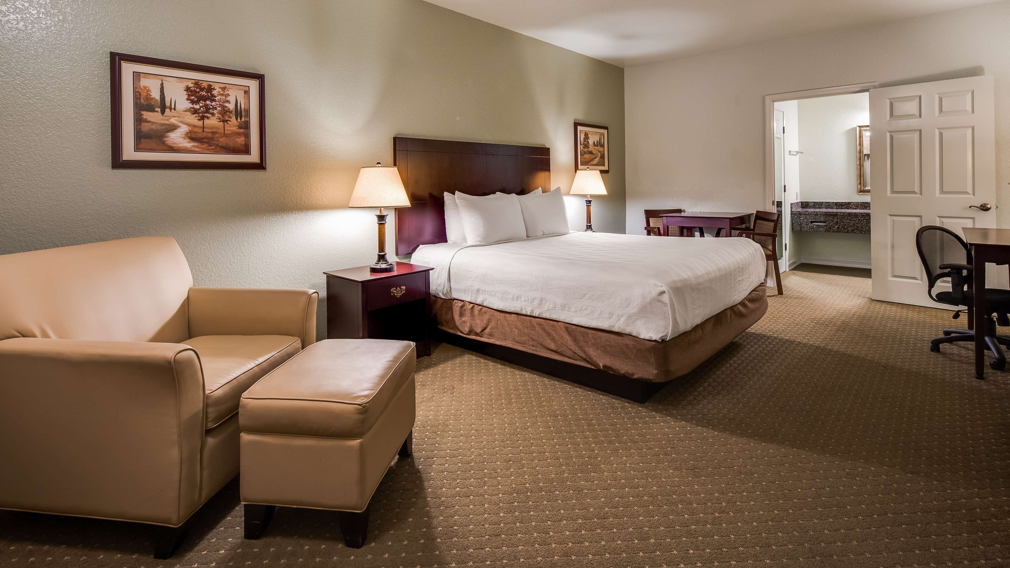 Una habitación de hotel con una cama, una silla, un reposapiés, una mesita de noche con lámpara y una puerta que da a otra habitación.